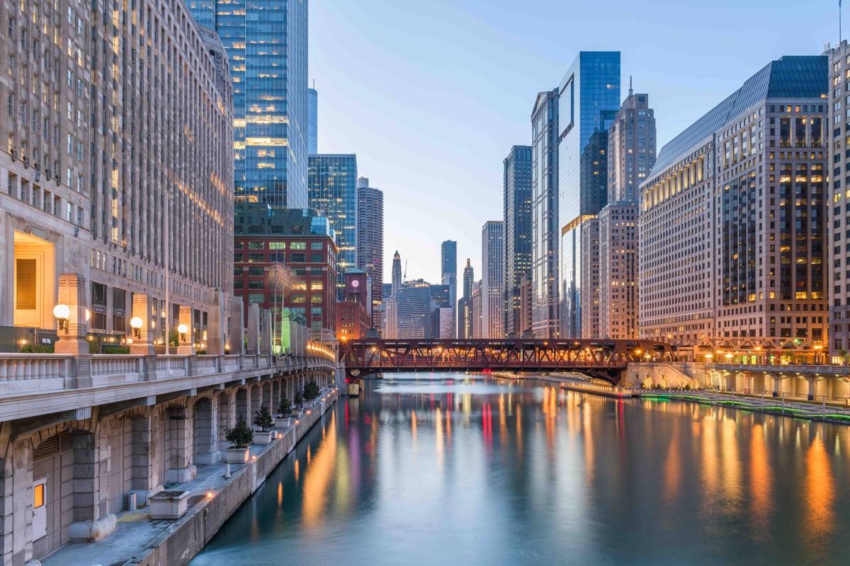 The Chicago River at dusk, framed by high-rise towers on both banks, with a bascule bridge and building lights reflecting on the water.